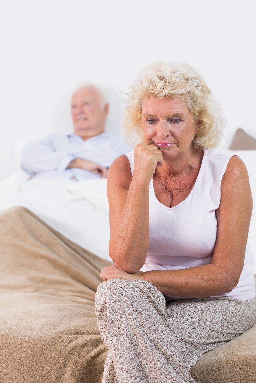 Discouraged old woman sitting on the bed with old man in background